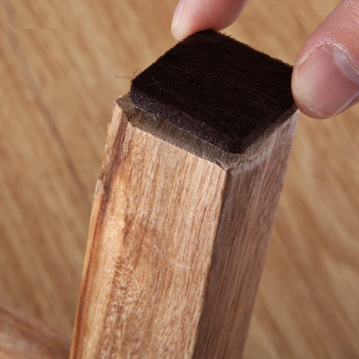 Square self-adhesive furniture pad for floor protection on a wooden surface.
