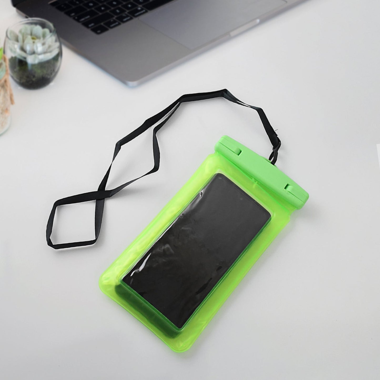A green clear phone pouch with a black smartphone and lanyard sits on a white desk with a silver laptop and small potted