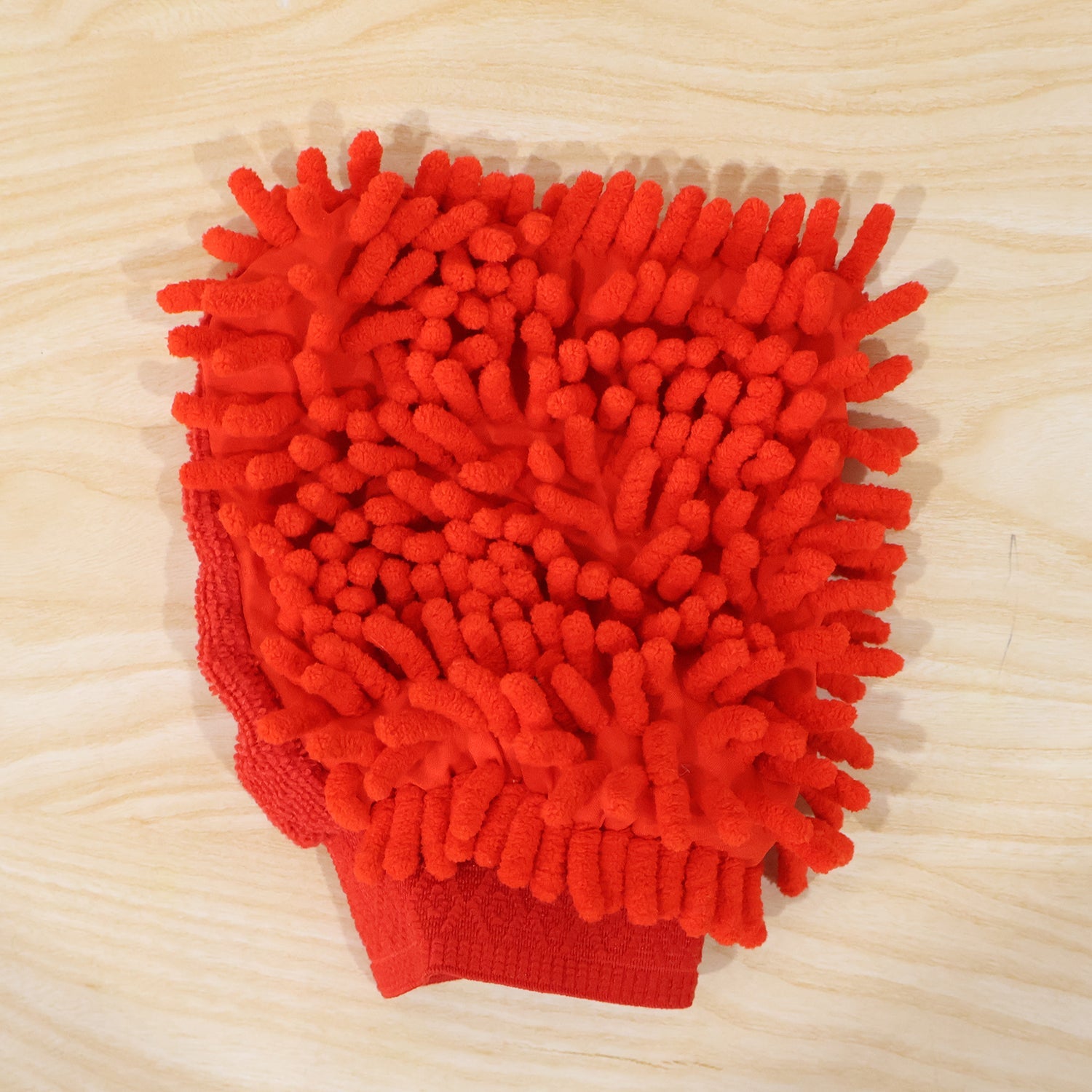 A red microfiber chenille wash glove, with a textured surface, rests on a light wooden surface.