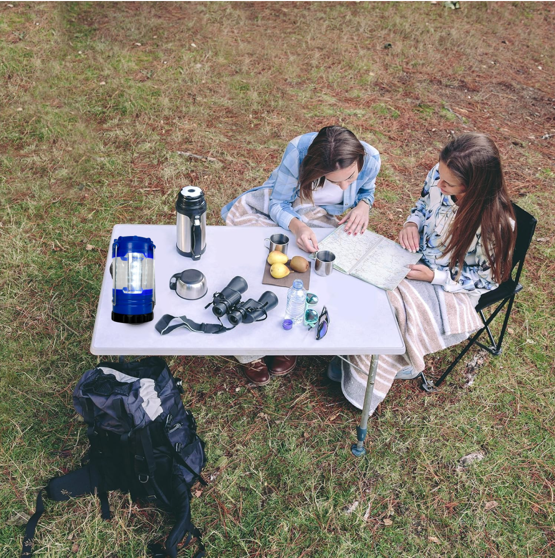 Two people sit at a white table outdoors, with a blue and black LED lantern, binoculars, and map on top.